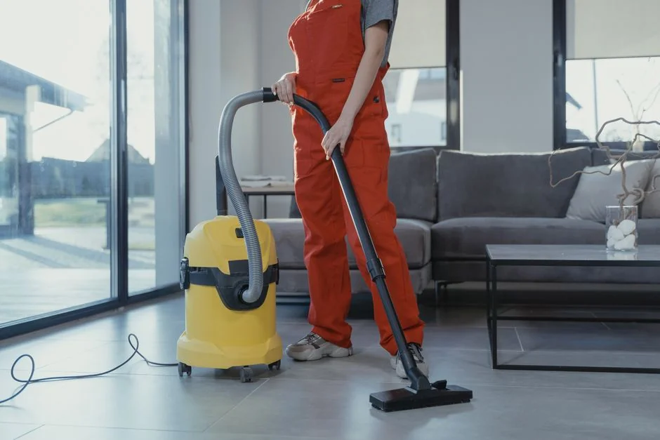 An adult woman in red coveralls operates a vacuum cleaner in a modern indoor setting, focusing on cleaning tasks.